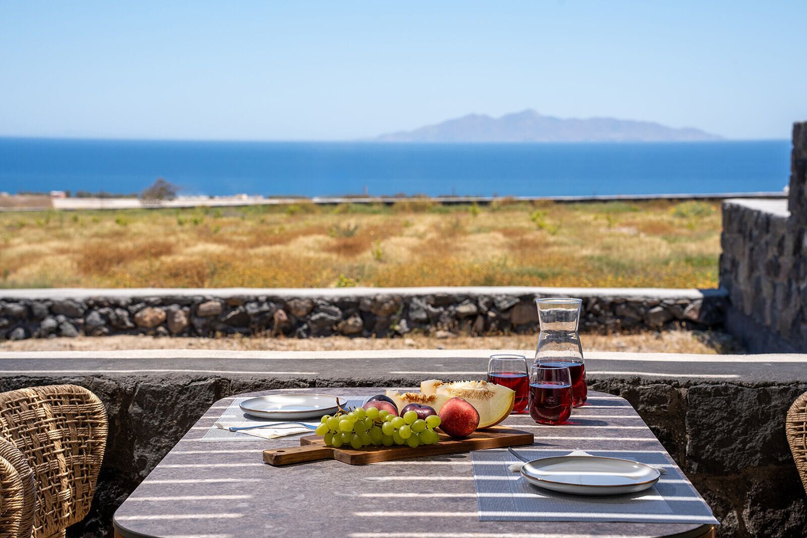 A table set with a plate, a bottle of red drink, a wooden board with grapes and fruit, and a view of the sea and mountains.