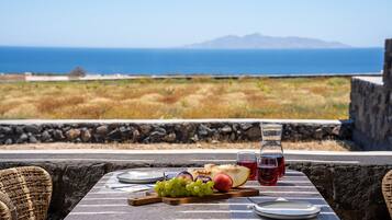 A table set with a plate, a bottle of red drink, a wooden board with grapes and fruit, and a view of the sea and mountains.
