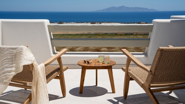 Un balcón con dos sillas de mimbre y una mesa redonda pequeña, con vista a una playa y montañas lejanas.