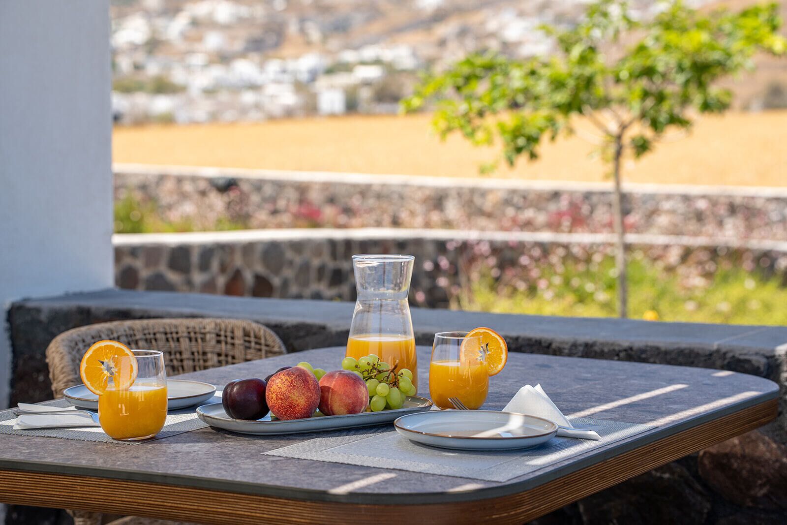 A table with a fruit platter, a pitcher of orange juice, and two glasses of orange juice on a balcony overlooking a scenic view.
