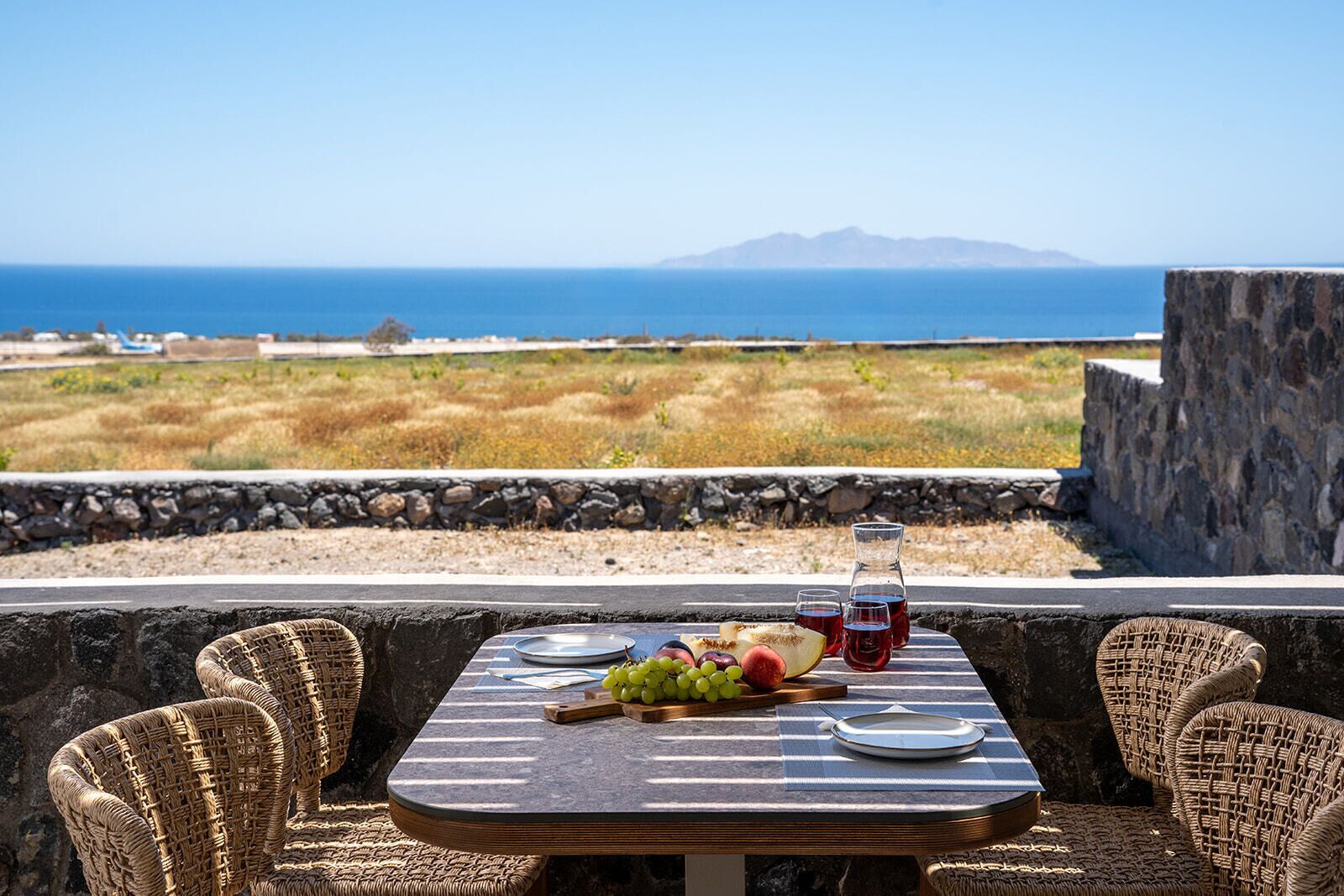 A table set outdoors with a view of the sea and mountains, wicker chairs, and a stone wall.