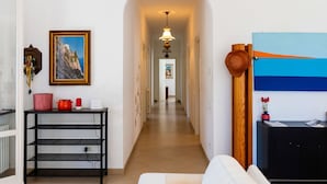 A hallway with a white wall, a framed painting, a shelf with red and white items, a black metal shelf, and a wooden door with a blue and orange artwork.