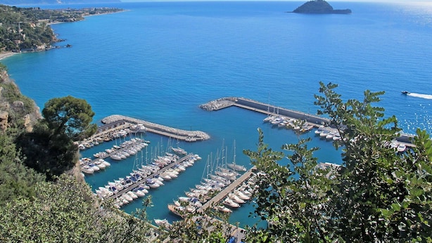 A marina with numerous boats docked, surrounded by a coastal landscape with hills and a clear blue sea.