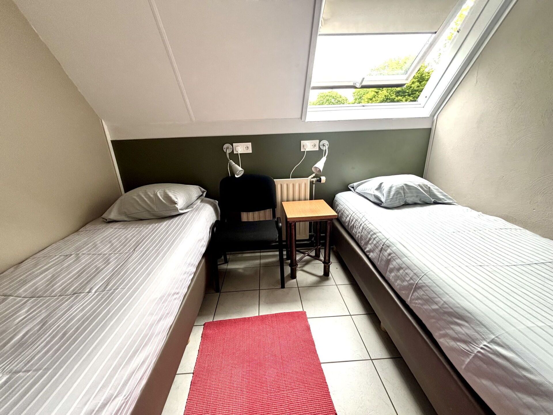 Een kleine, compacte kamer met twee eenpersoonsbedden, een bureau en een stoel. Er is een raam dat natuurlijk licht en uitzicht op groen biedt.