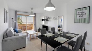 Un salon moderne comprenant un coin repas, un canapé, une table dressée pour quatre personnes et un panneau indiquant qu’un club propose des petits-déjeuners le dimanche.