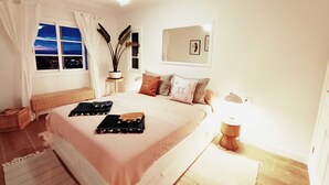 Ein Schlafzimmer mit einem großen Bett, einem Nachttisch, einer Topfpflanze und einem Fenster mit Blick auf die Stadt.