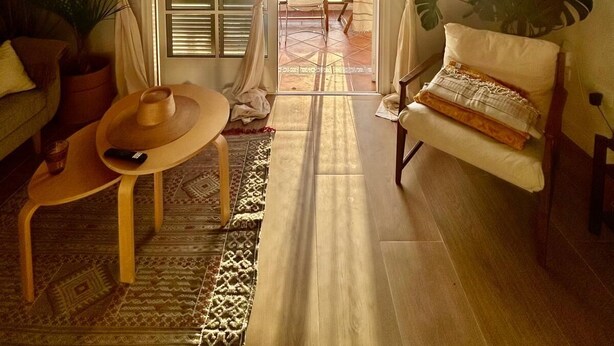 Ein gemütliches Wohnzimmer mit Sofa, Sessel, kleinem Tisch und einer Pendelleuchte. Eine Glastür öffnet sich zu einer Außenpatio-Terrasse.
