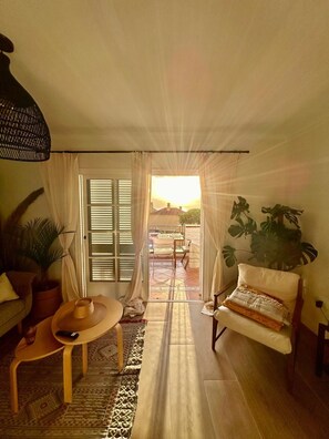 Un salón acogedor con un sofá, una silla, una mesita y una lámpara colgante. Hay un ventanal corredero que da a un patio exterior.