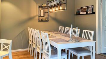A dining room with a long table, white chairs, a modern pendant light, and framed pictures on the wall.