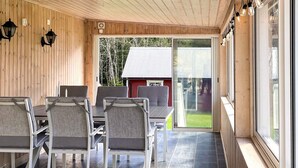 A covered patio area with a table and chairs, wooden walls, and a view of a red building through a glass door.