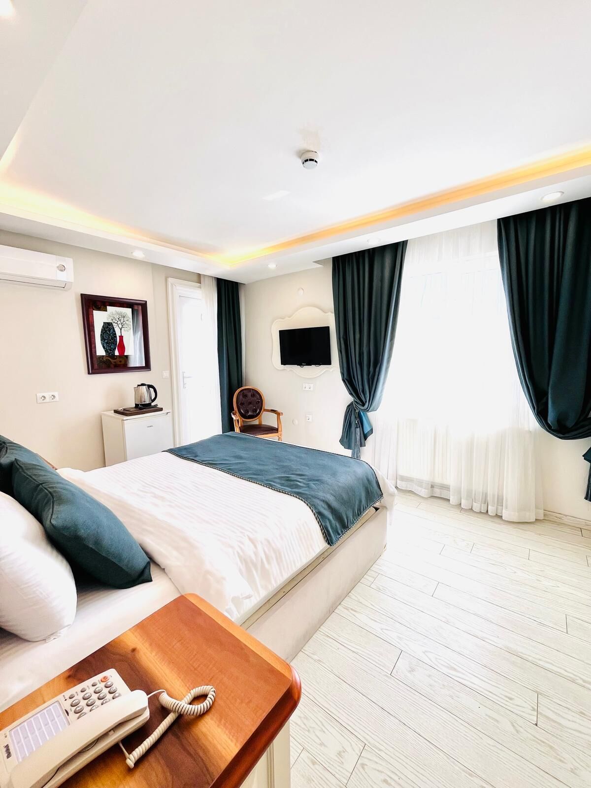 A hotel room with a bed, a bedside table with a phone, a TV on the wall, and a window with curtains.