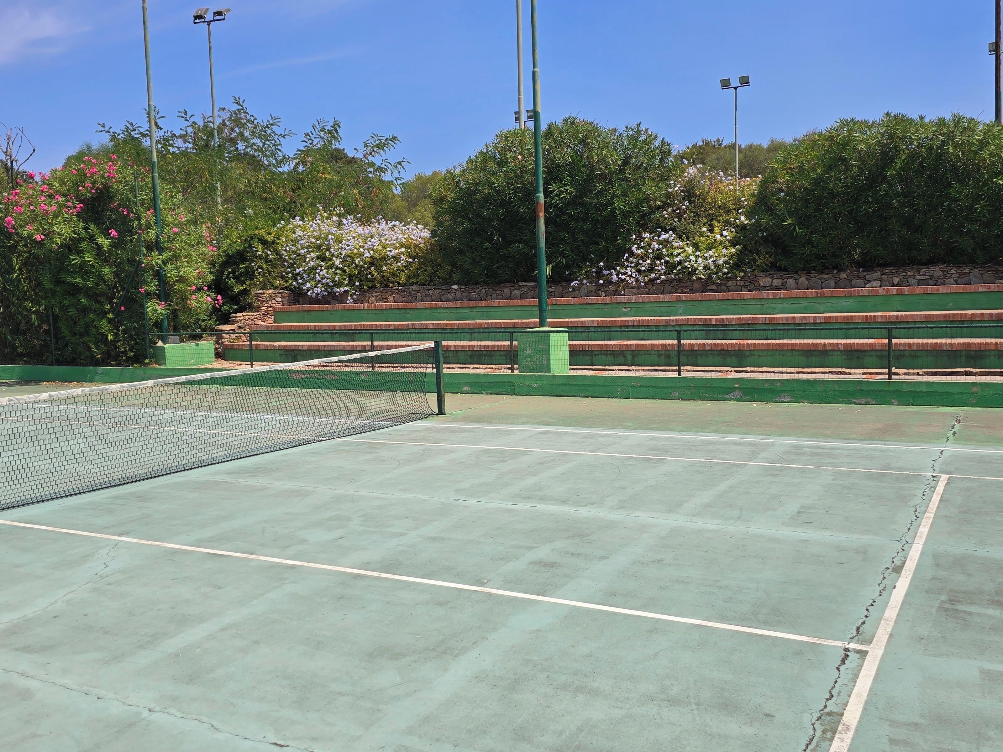 A tennis court with a net, surrounded by green seating and trees with pink flowers.