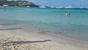 Une plage de sable fin bordée d’une eau turquoise et limpide, avec des bateaux et, au loin, une colline parsemée de bâtiments.