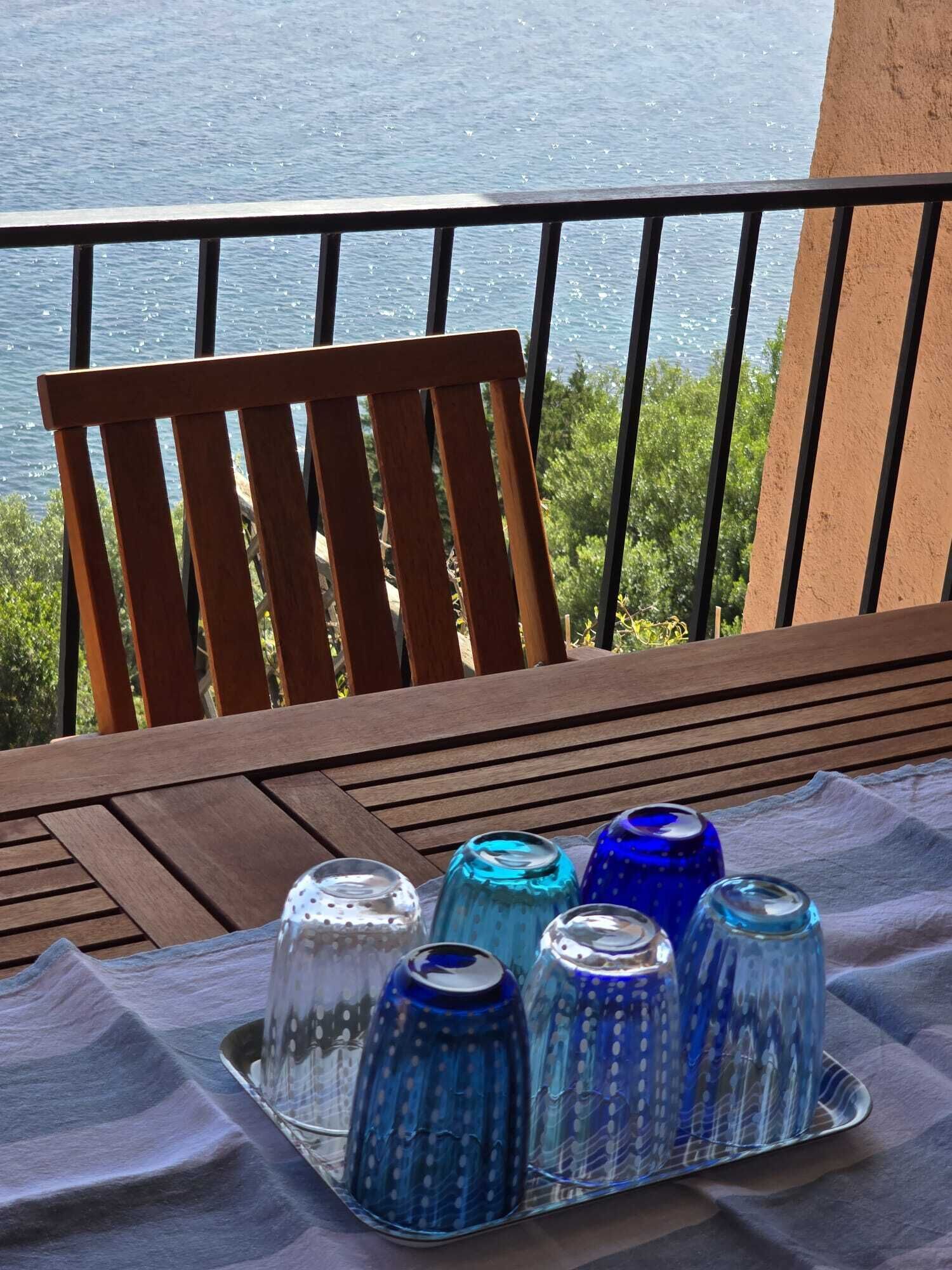 A wooden chair and a table with a tray of decorative glassware on a balcony overlooking a body of water.
