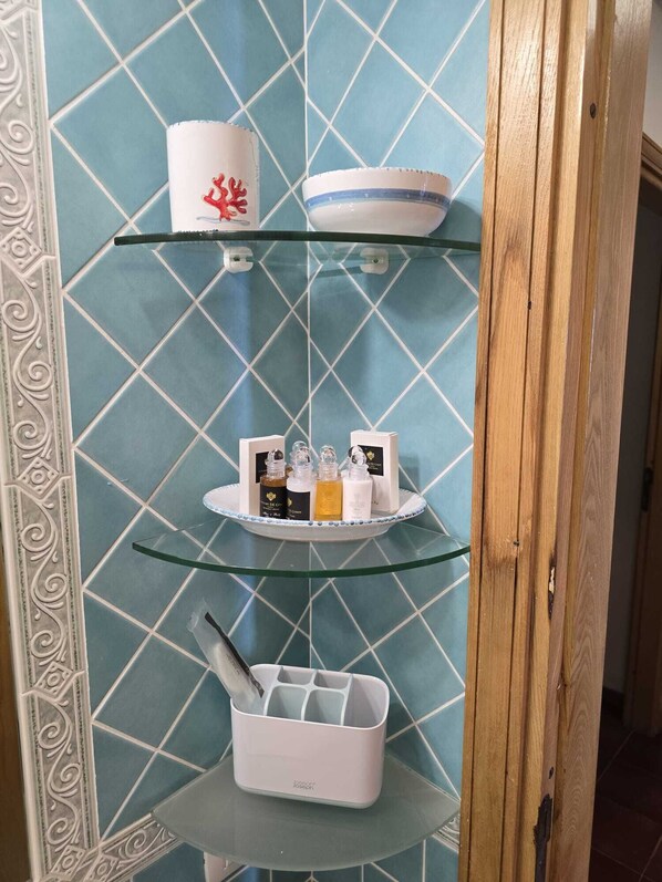 A bathroom with a blue tiled wall, a white decorative border, and a glass shelf unit holding toiletries.