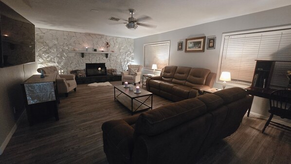 A living room with a stone fireplace, a ceiling fan, and a large window with blinds. - 3-bedroom with Lakeview, Free Kayaks and Game Room, Pets Allowed, Lanai & Grill (Inverness)