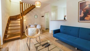Ein modernes Wohnzimmer mit einem blauen Sofa, einem Korbstuhl, einer Holztreppe und einem Essbereich mit Tisch und Stühlen.