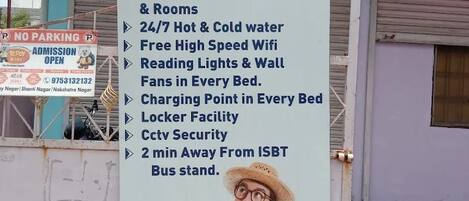 A signboard for a 5-star dormitory listing amenities such as AC/Non AC dormitory, 24/7 hot & cold water, free high-speed wifi, reading lights, wall fans, charging points, locker facility, CCTV security, and proximity to ISBT bus stand.