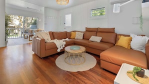 A modern living room with a brown sectional sofa, a round coffee table, and a large window providing a view of the outdoors. - The Point Arkwright Bungalow (Point Arkwright)