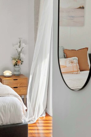 Ein Schlafzimmer mit einem Bett, einem Nachttisch mit Lampe, einer Vase mit Blumen und einem Spiegel, der den Raum reflektiert.
