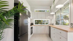 A modern kitchen with white cabinets, a stainless steel refrigerator, and a window providing a view of greenery outside. - The Point Arkwright Bungalow (Point Arkwright)