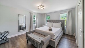 A bedroom with a large bed, a bench, a mirror, and windows with curtains.