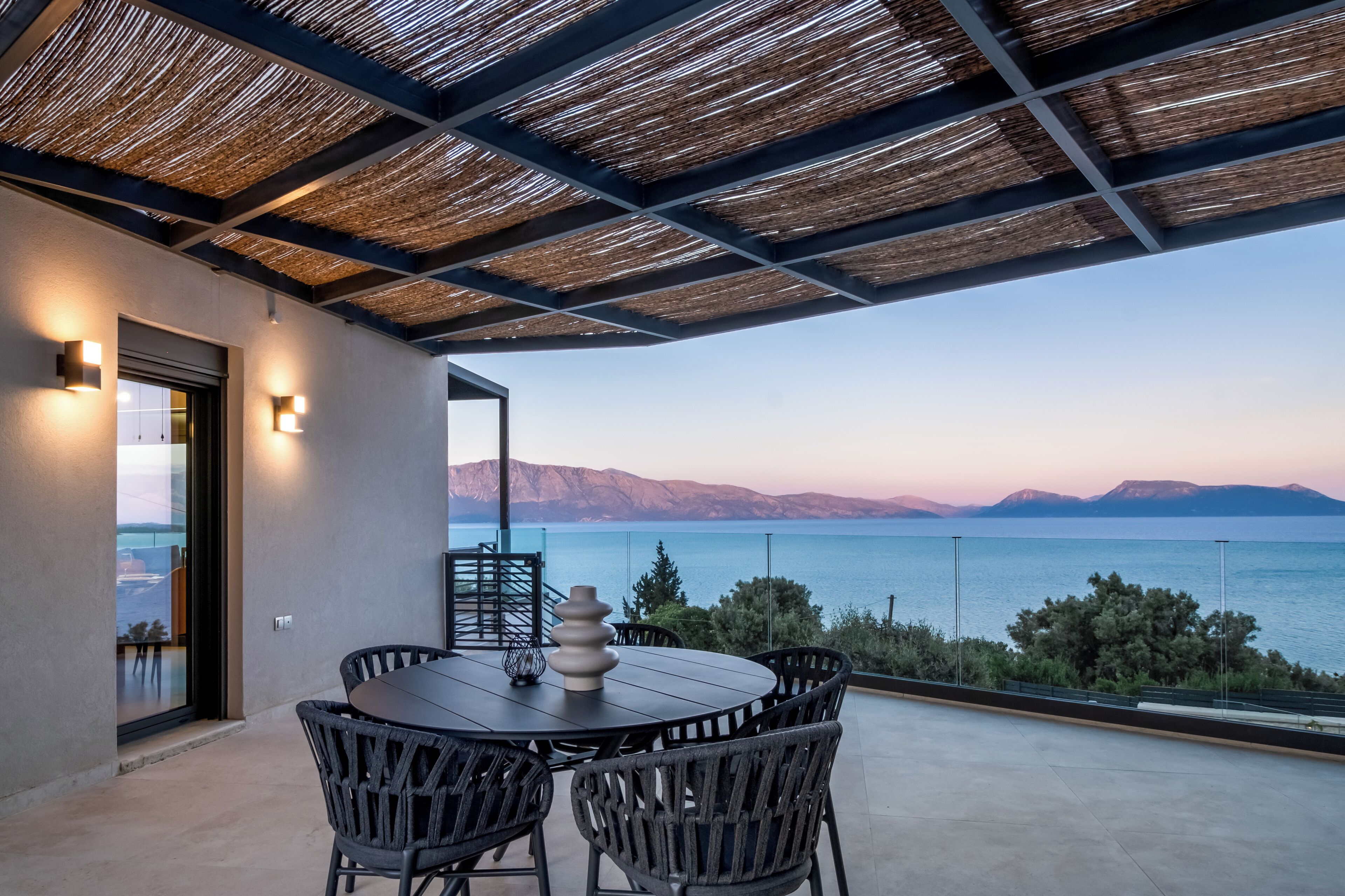 A terrace with a round table and chairs, a glass door leading to a balcony, and a view of the sea and mountains.