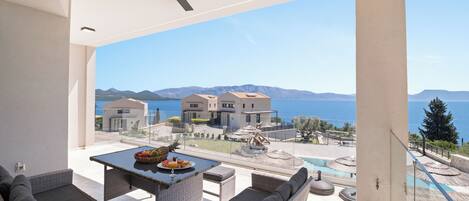 A balcony with a ceiling fan, a table with fruit and a glass, and a view of a pool and buildings across the water.