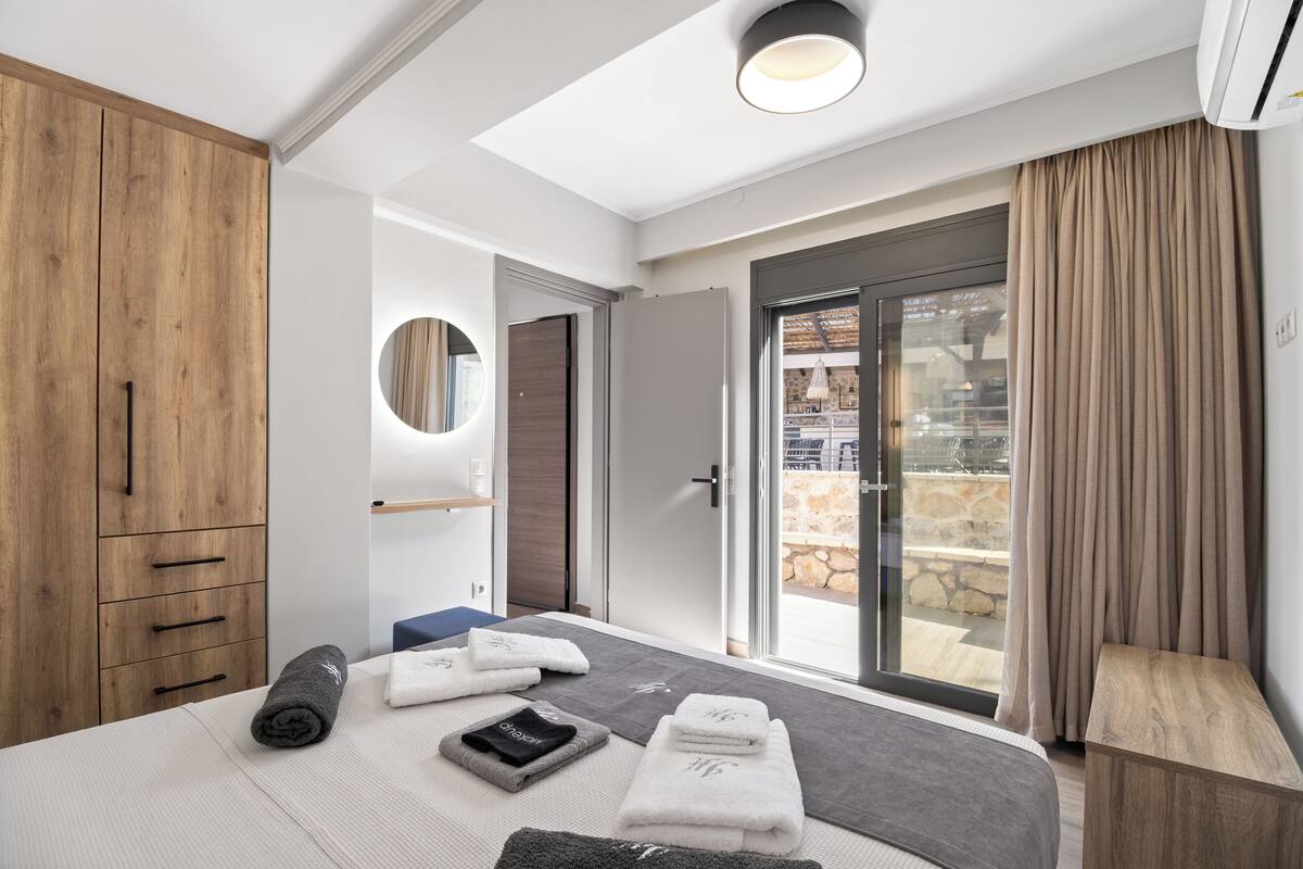 Une chambre d’hôtel moderne avec un grand lit, une armoire en bois, un miroir rond et une vue sur l’extérieur grâce à une porte vitrée.