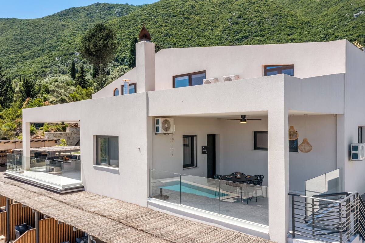 Une villa moderne dotée d’un balcon en verre, d’une piscine et offrant une vue sur une montagne verdoyante.