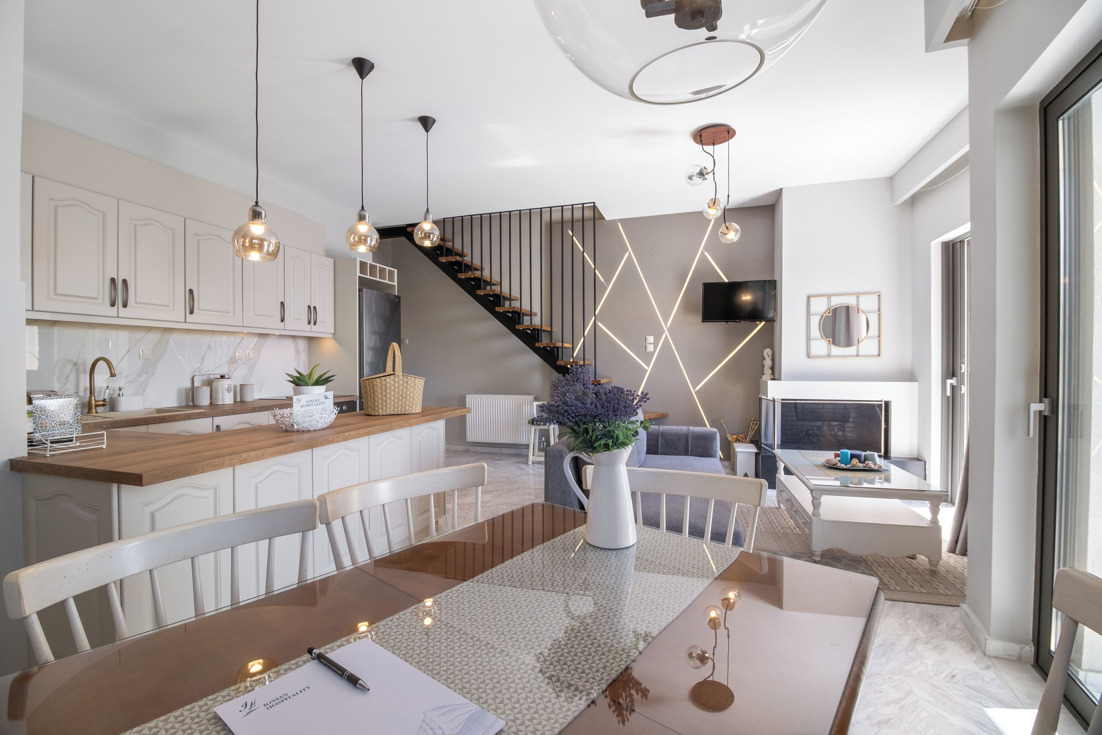 Una cocina moderna con armarios blancos, encimera de madera e iluminación colgante. Hay una zona de comedor con una mesa y sillas, y una escalera que sube a otro nivel.