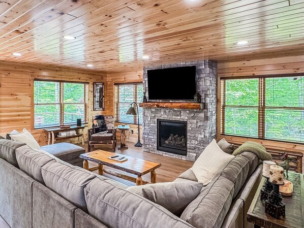 A log cabin interior with a stone fireplace, a television mounted above it, and a large window providing a view of the outdoors.