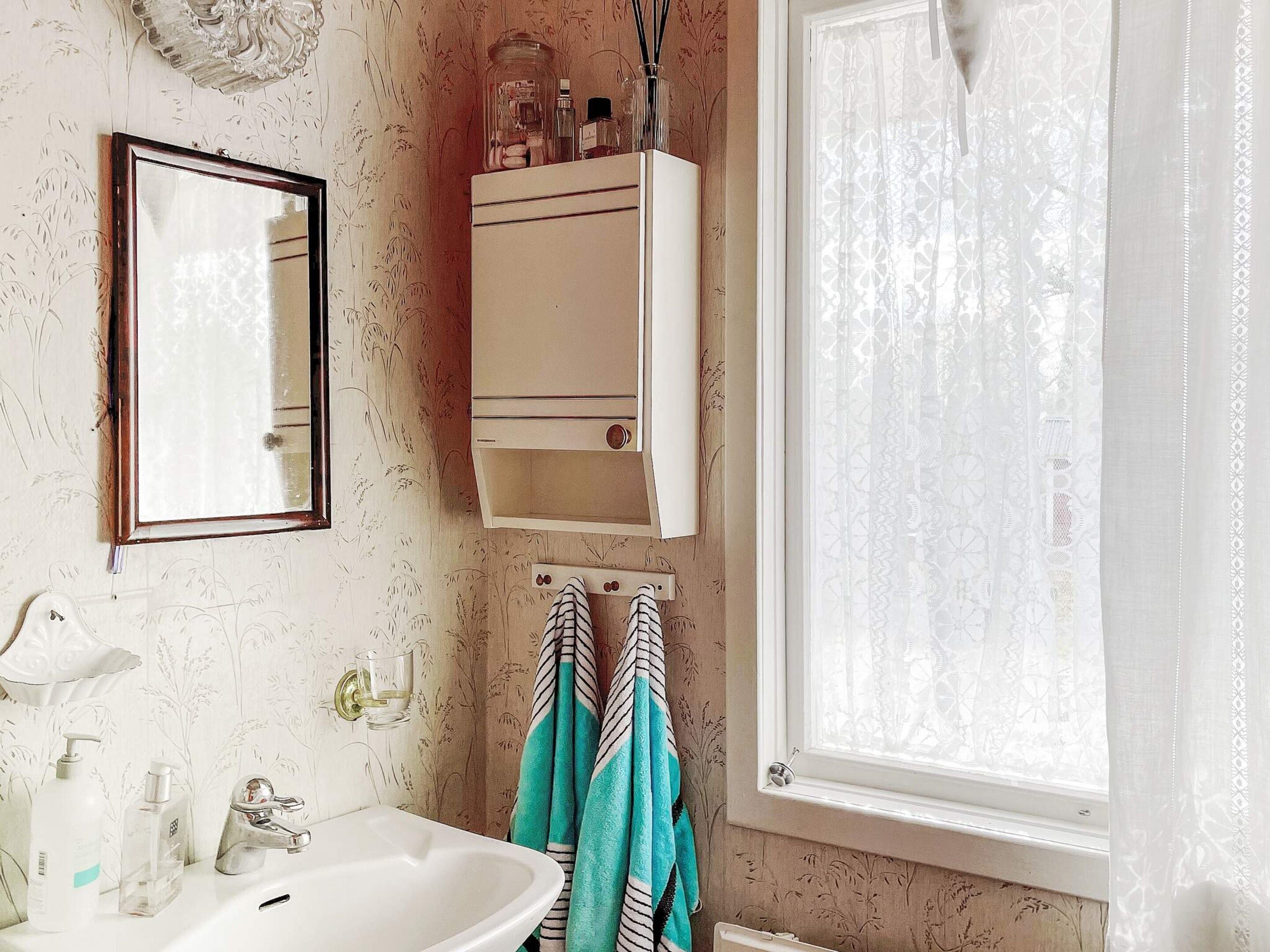Ein Badezimmer mit einem weißen Waschbecken, einem Spiegel, einem Schrank mit Pflegeprodukten und einem Fenster mit Vorhang.