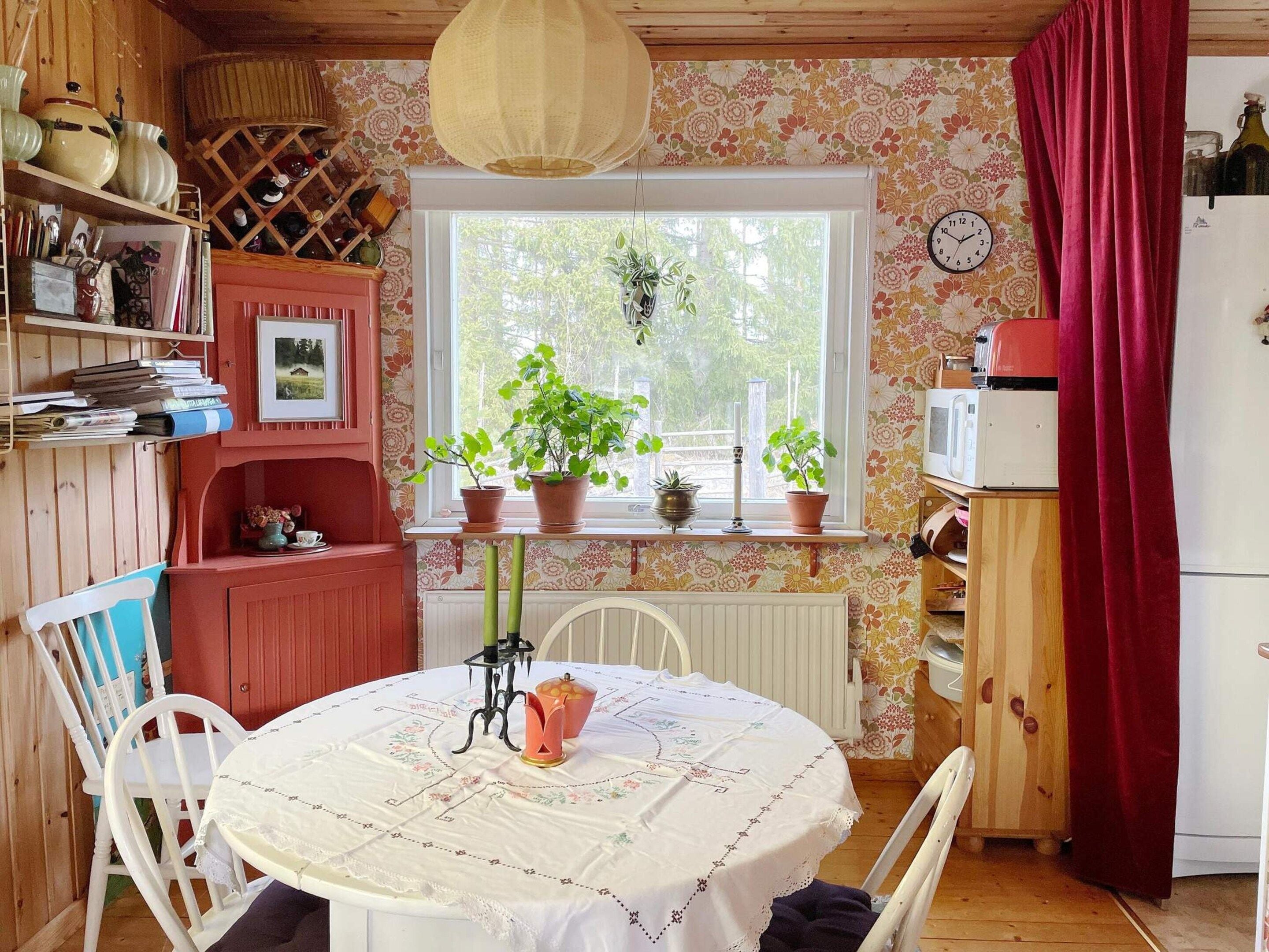 A cozy kitchen with wooden cabinets, a dining table with chairs, a window with plants, and a clock on the wall.