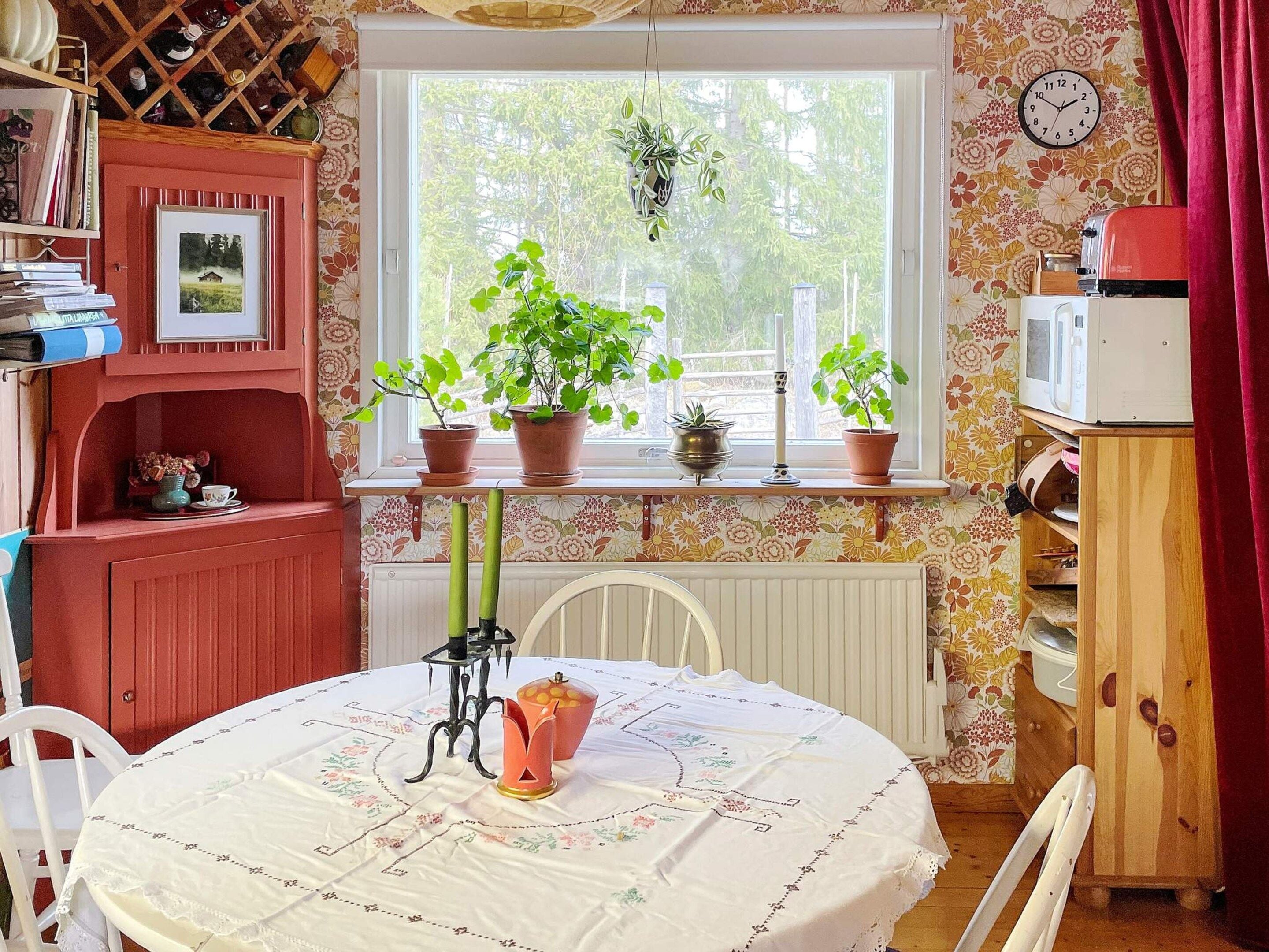 Una habitación acogedora con una mesa redonda, un armario rojo, un reloj en la pared y varias plantas en macetas en el alféizar de una ventana.