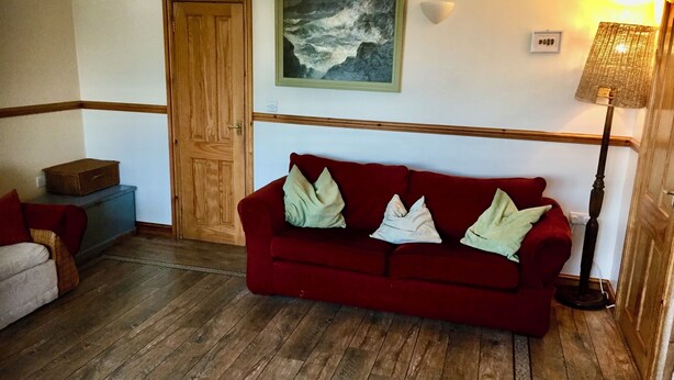 Une pièce avec un plancher en bois, un canapé rouge avec des coussins, une porte en bois, un tableau encadré au mur, une lampe et une petite table sur laquelle se trouve une boîte.
