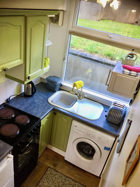 A compact kitchen with a stove, sink, and washing machine. - Rustic Apartment with Sea views, and a short walk to the beach. (Freshwater East)