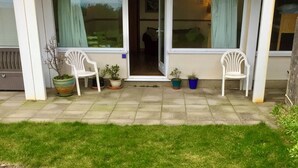 Une terrasse couverte avec deux chaises en plastique blanches, des plantes en pot et une porte vitrée donnant sur un espace intérieur.