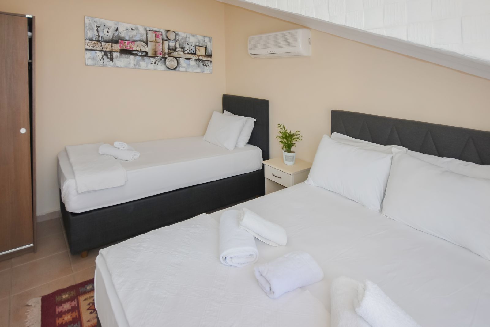 A hotel room with two beds, a painting on the wall, an air conditioner, and a small plant on the nightstand.