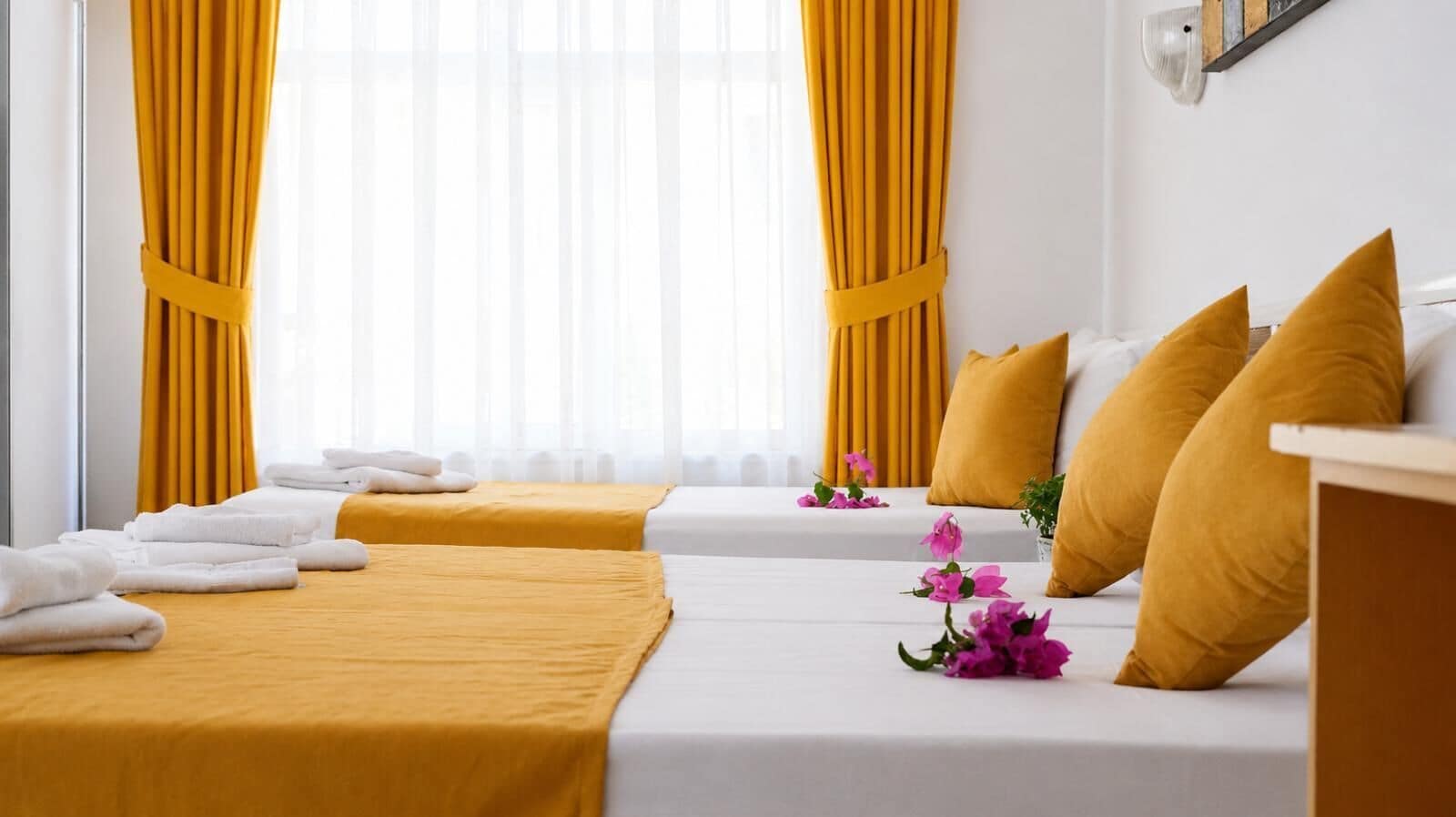 Habitación de hotel con dos camas, cortinas amarillas, ropa de cama blanca y una planta pequeña.