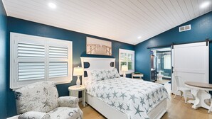 Een slaapkamer met een bed, twee nachtkastjes, een stoel, een tafel en een raam met jaloezieën.