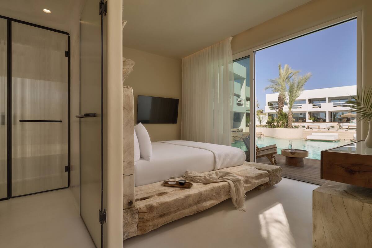 A modern hotel room with a large bed, a TV, and a view of a pool and building.