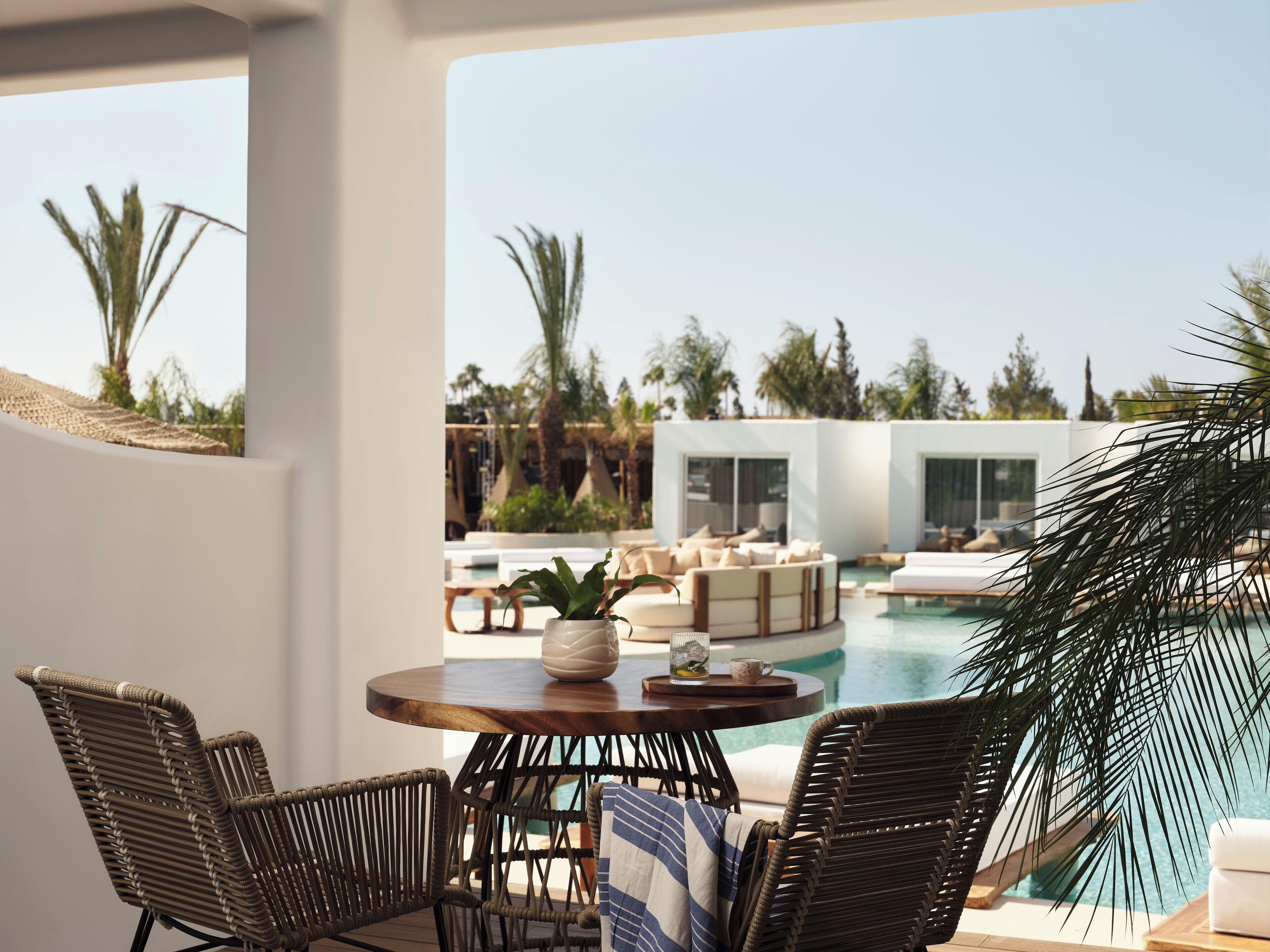 A poolside area with wicker chairs, a round table, and a view of a pool and modern buildings in the background.