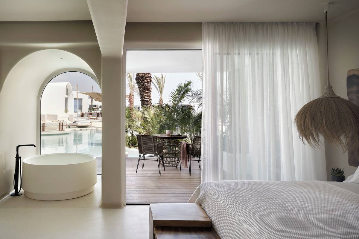 A modern hotel room with a large bed, a circular seating area, and a view of a pool and outdoor seating area.