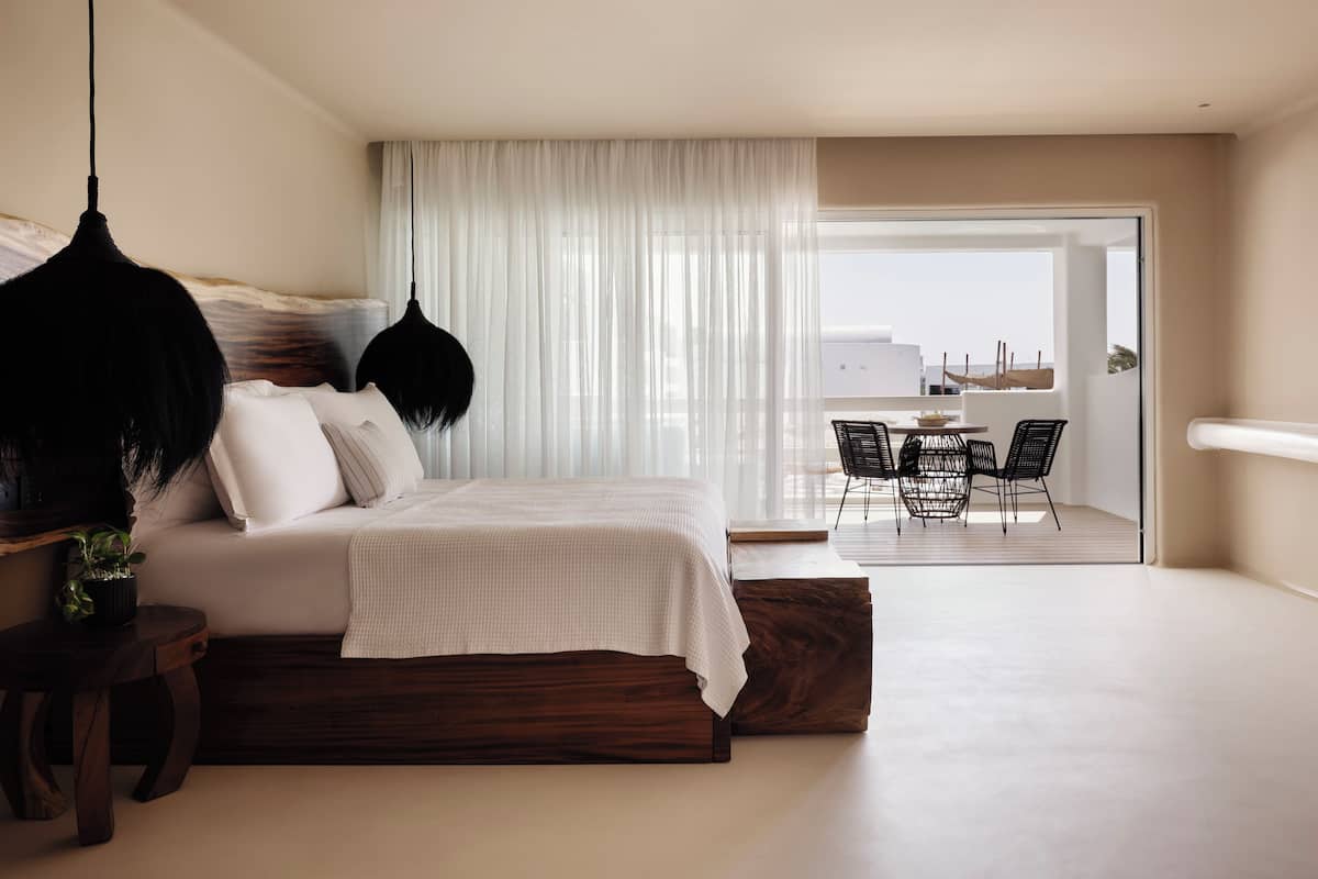 A modern bedroom with a large bed, a hanging light fixture, and a view of an outdoor balcony with chairs.
