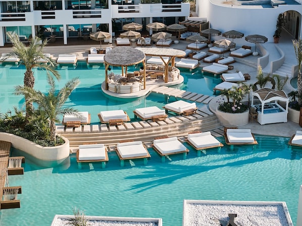 Zona de piscina en un complejo turístico con múltiples sillas de descanso, un área de estar central y un bar junto a la piscina.