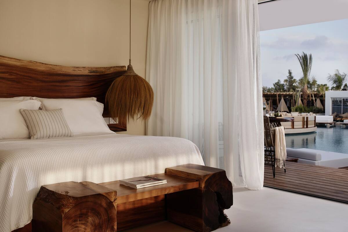 A bedroom with a large bed, a wooden headboard, a hanging light fixture, and a view of a pool and outdoor seating area.