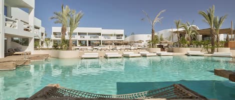 Zona de piscina con sillones de descanso y sombrillas, rodeada de palmeras y edificios modernos.