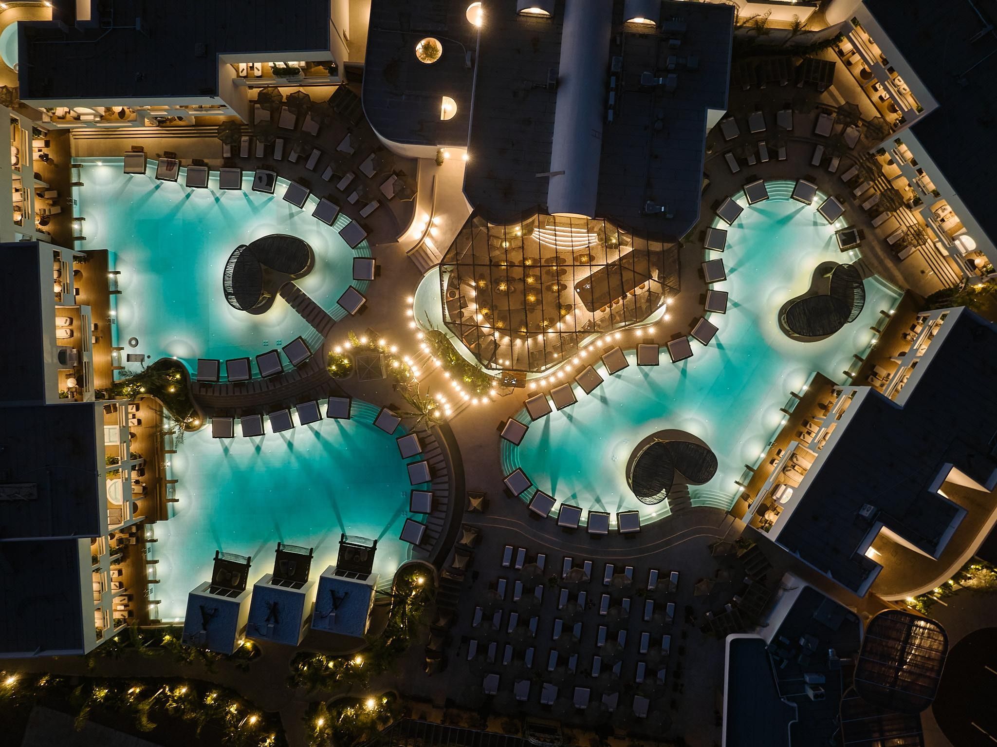 A rooftop pool area with multiple swimming pools, sun loungers, and a central seating area.