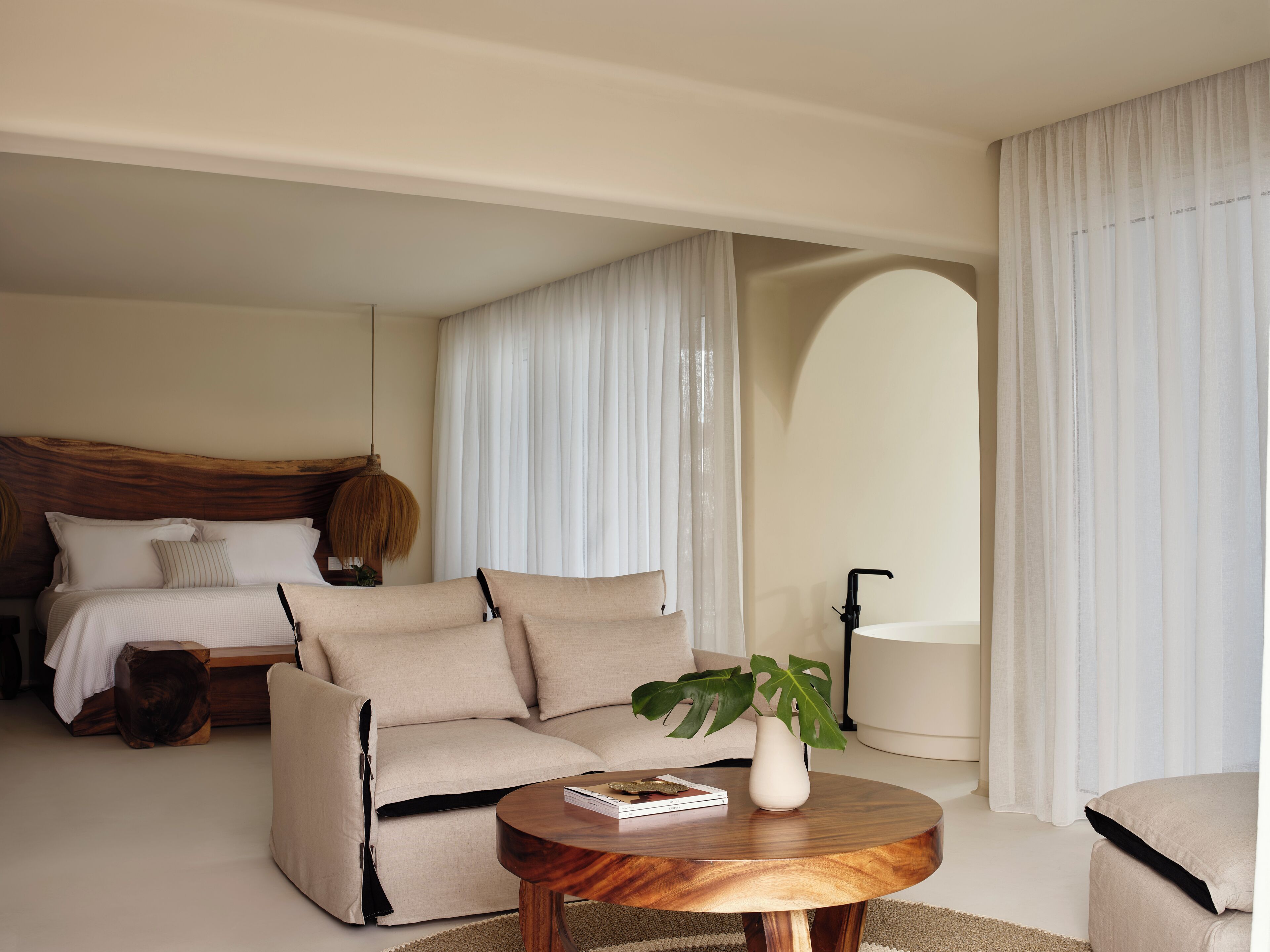 A modern bedroom with a bed, sofa, and a round wooden table with a book and a plant.